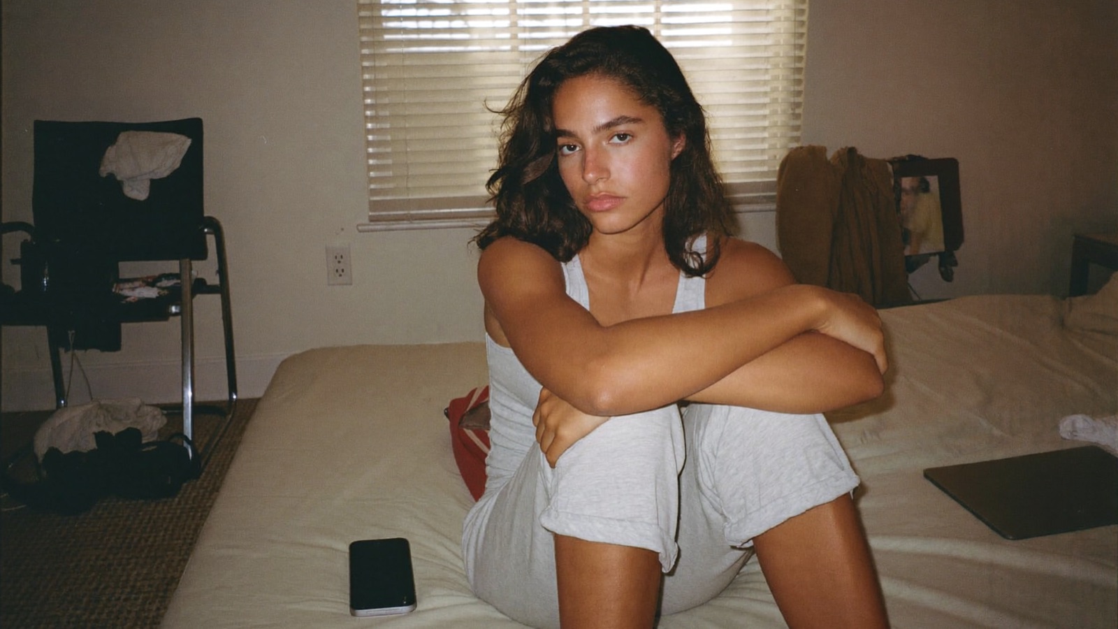 A young woman sitting on the edge of an unmade bed with her phone face down beside her, an account she stopped posting to